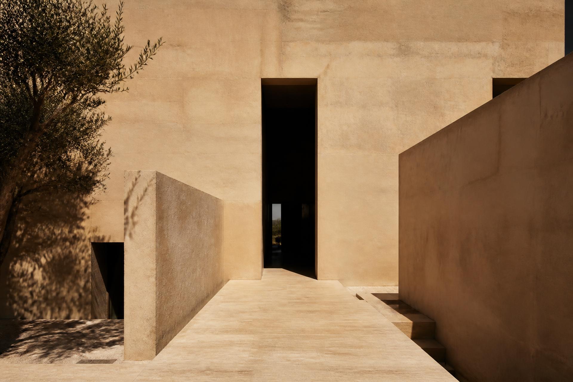 Monolithic sand-coloured concrete house with a tall dark central doorway in Mediterranean light